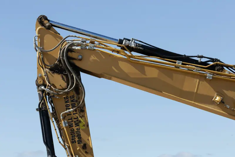 The yellow boom and dipper arm of an excavator against a blue sky, hydraulic hoses along the arm