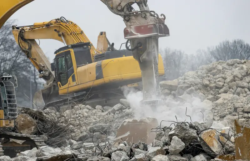 An excavator with a hydraulic breaker works through broken concrete and rebar on a demolition site