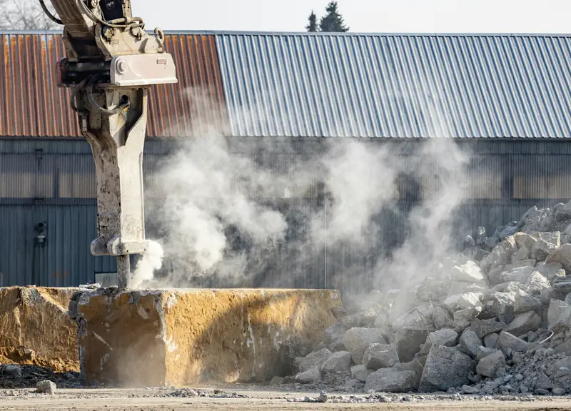 A hydraulic breaker on an excavator arm cracks a thick concrete slab, dust rising beside a pile of broken concrete