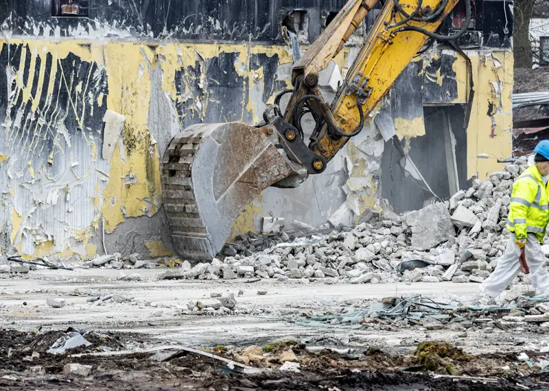 An excavator with a demolition bucket pulls down a rendered wall while an operative in hi-vis walks clear of the work area