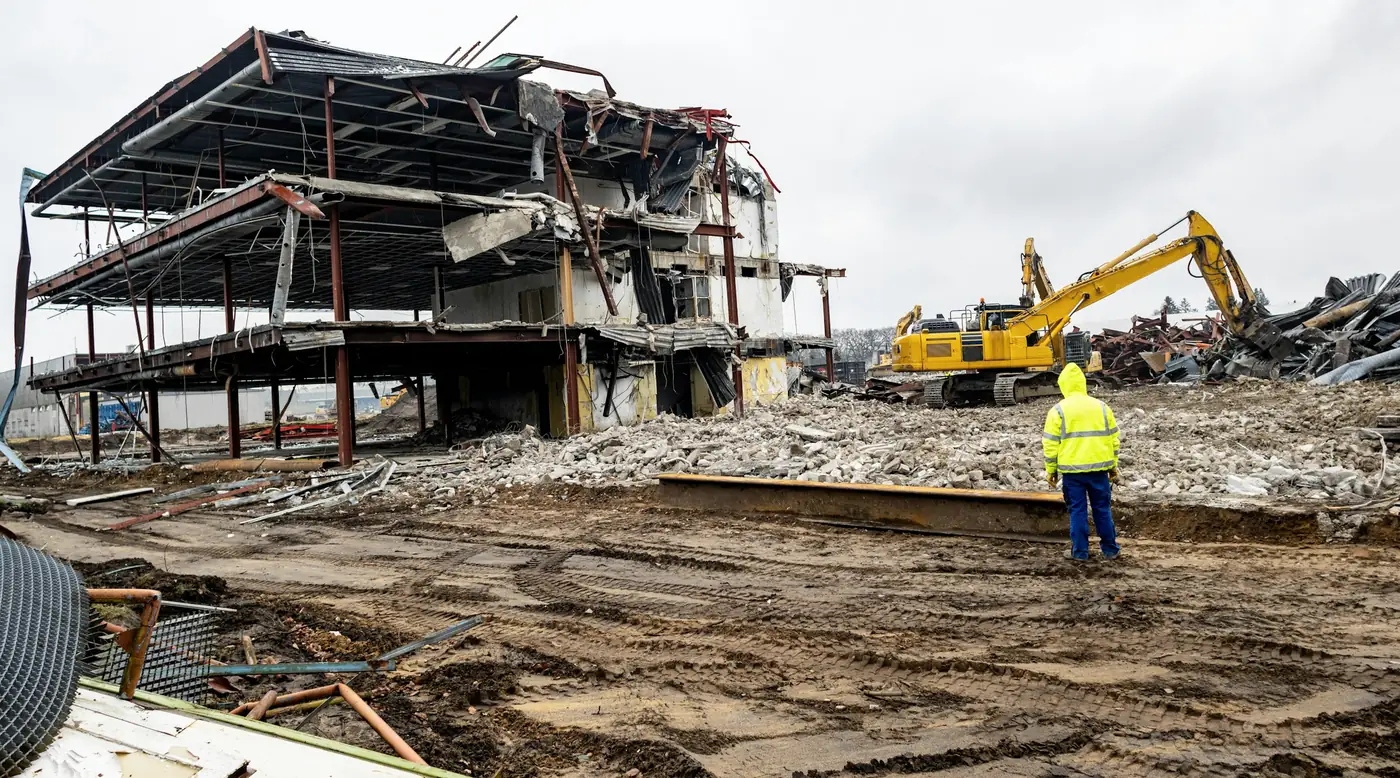A cleared demolition site: an operative in hi-vis stands on levelled ground, the last steel frame being taken down behind him by an excavator