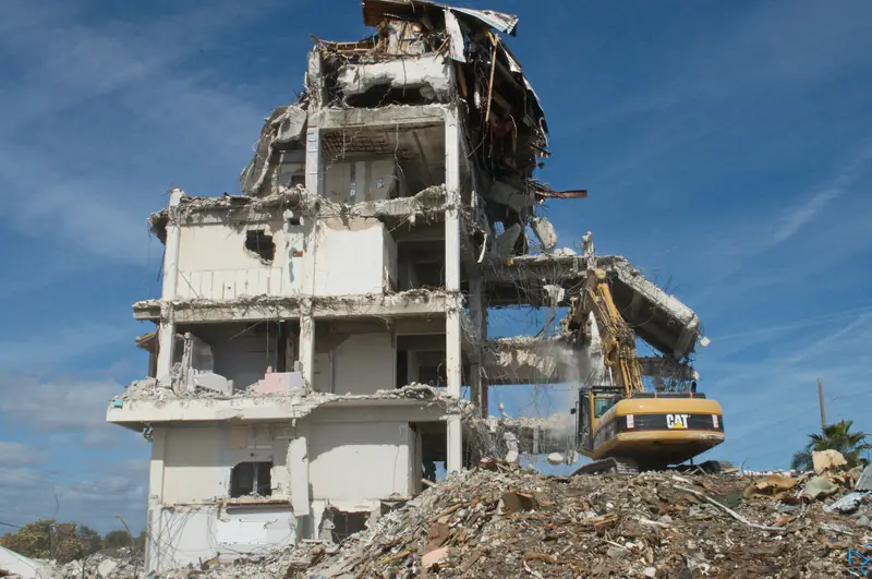 An excavator working on top of a rubble pile takes down a multi-storey concrete frame, floors exposed