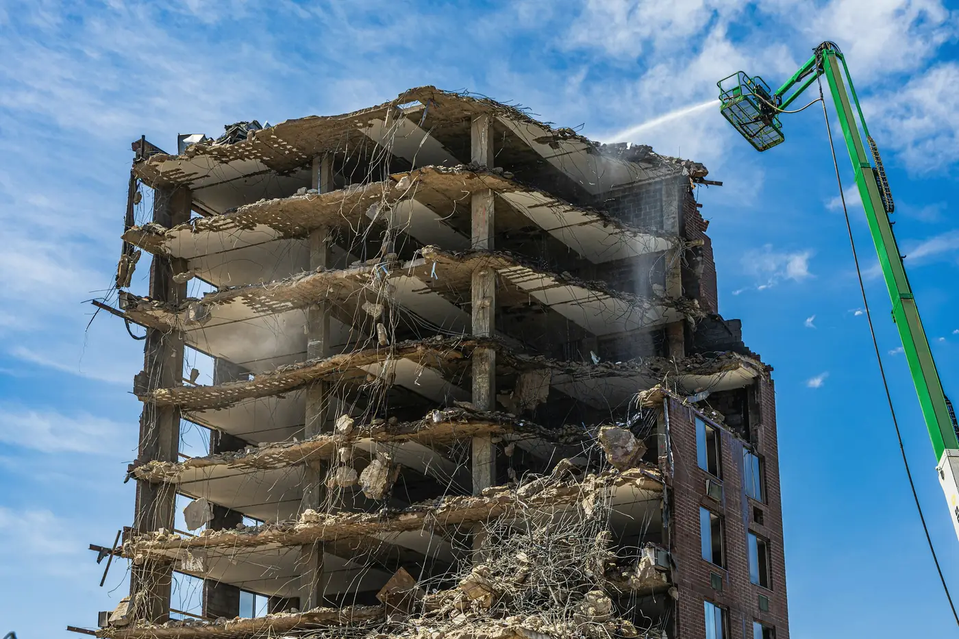 A concrete-frame block half taken down, floors stripped to the slab, while a high-reach boom sprays water to keep the dust down