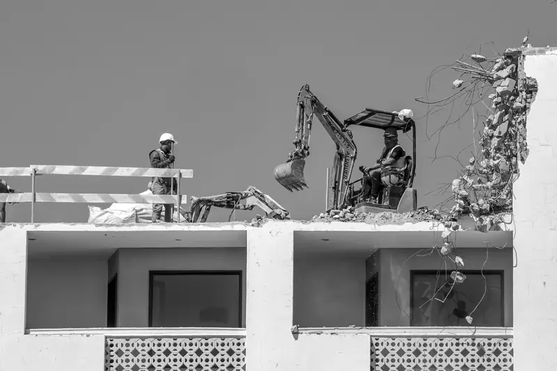 Black-and-white photo: two compact excavators and an operative working on the roof slab of a block, taking it down from the top