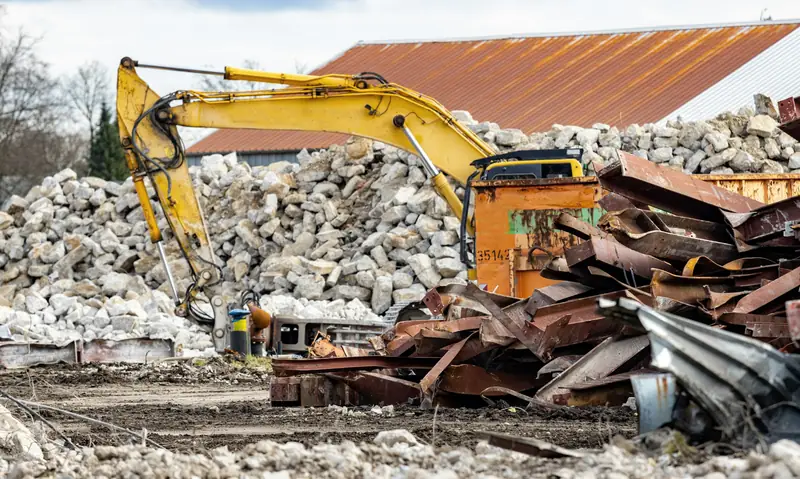 Sorted stockpiles on a cleared site: crushed concrete behind, recovered steel sections in front, an excavator between them